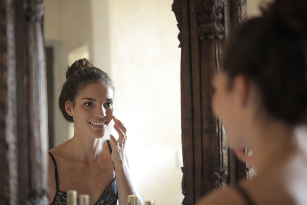 our-mission Young woman applying skincare and smiling at her reflection in a rustic mirror.